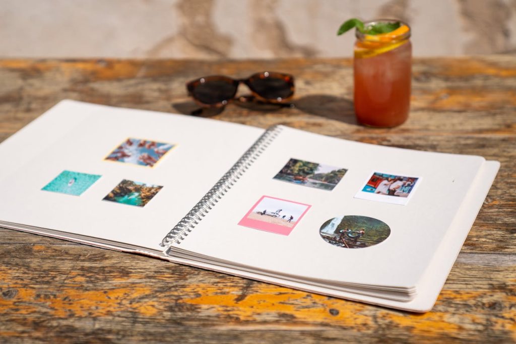Open photo album on a wooden table with sunglasses and drink, evoking summer memories.